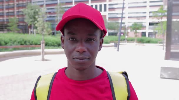 Repartidor afroamericano con gorra roja y camiseta sonriendo a la cámara — Vídeos de Stock