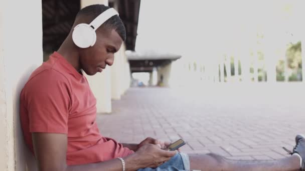 Vue latérale du jeune homme afro-américain avec écouteurs utilisant un téléphone mobile — Video