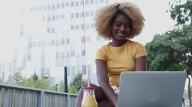 Genç siyahi Amerikalı kadın dışarıda bankta otururken dizüstü bilgisayar kullanıyor.