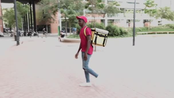 Zwarte bezorger in rood uniform met rugzak die binnenkomt kantoorgebouw — Stockvideo