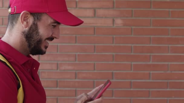 Side view of happy hispanic delivery man in red clothes using mobile phone — Stock Video