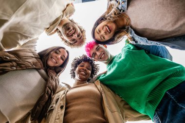 Smiling young group of people standing together in circle at city street. Low angle view of millennial trendy student friends laughing at camera