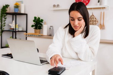 Young woman wearing bathrobe calculating domestic expenses using calculator and laptop in kitchen at home