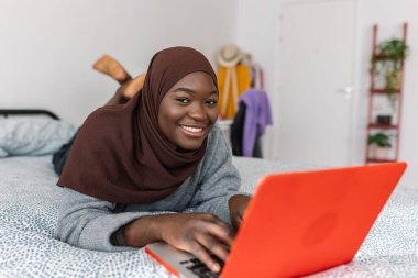 Portrait of young african woman working on laptop while smiling at camera relaxed on bedroom. Technology and education concept.