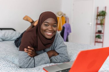 Joyful portrait of young teen african girl in muslim headscarf with laptop computer smiling at camera while lying on bed at home. Technology and education concept.