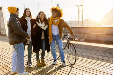 Group of young multi-ethnic friends having fun together outdoors during wintertime at the port, one of them is holding a bicycle