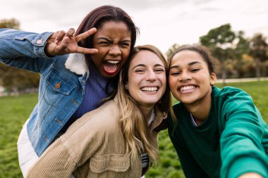 Happy multiracial college student friends enjoying outdoor time together, taking a fun close-up selfie in a park