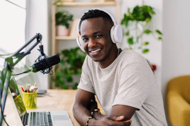 Portrait of young happy African student man wearing headphones and using a professional microphone, creating digital content for his podcast