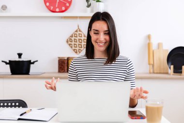 Gülümseyen kadın serbest çalışan, uzaktan çalışan, bilgisayarında mutfaktaki bir video görüşmesi olan. İş ve eğitim kavramı