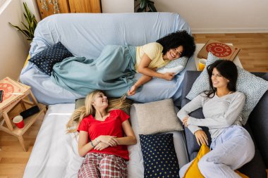 Üç neşeli genç kız pijama partisinin tadını çıkarıyor, kanepede ve yerde dinleniyor, sohbet ediyor ve eğleniyorlar. Kadın arkadaşlığı ve gençlik kavramı