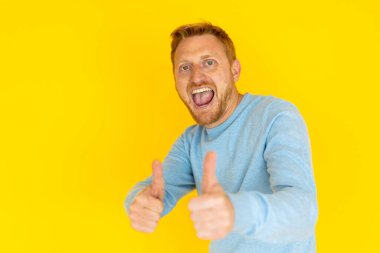 Heyecanlı kızıl saçlı adam iki eliyle baş parmağını kaldırıyor, pozitif olduğunu ve sarı renkli arka planda onay verdiğini söylüyor.