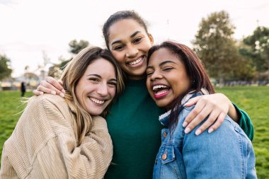Üniversite kampüsünün yeşil bölgesinde kameraya mutlu bir şekilde gülümserken üç bayan arkadaş birbirlerine sarılıyorlar. Çok kültürlü gençlik, arkadaşlık ve öğrenci yaşam tarzı konsepti