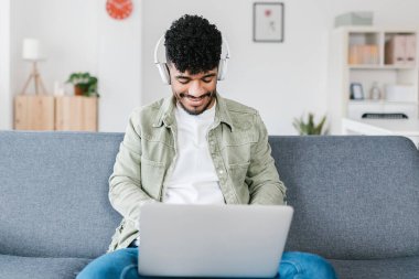 Kablosuz kulaklıkla müzik dinleyen genç adam evdeki koltukta otururken dizüstü bilgisayarla çalışıyor.
