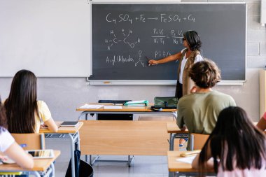 Bayan öğretmen lise öğrencilerine tahtadaki kimya formüllerini açıklıyor. Eğitimci modern bir sınıfta bilim öğretiyor ve öğrencilerle iletişim kuruyor. Bilim ve eğitim kavramı.