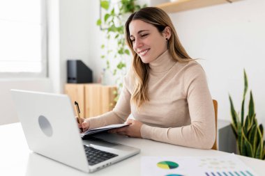 Gülümseyen iş kadını evinden bilgisayarına çalışırken not defterine not alıyor. Bitkiler ve ışıkla dolu parlak modern çalışma alanı. Verimlilik, odaklanma ve iş-yaşam dengesi kavramı.