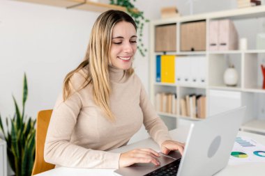 Masasına gülümserken dizüstü bilgisayarında yazan beyaz bir iş kadını. Parlak bir alanda çizelgeler ve belgeler ile modern uzaktan kumanda kurulumu. Dijital iş başarısı kavramı.