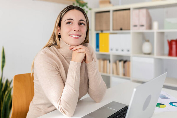 Расслабленная молодая деловая женщина сидит за своим столом, улыбаясь и глядя в сторону от экрана. Современное и уютное домашнее рабочее место с заводами и книжными полками. Концепция счастливого образа жизни на дому.