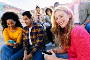 Bir grup çok ırklı öğrenci dışarıda akıllı telefonu paylaşırken, genç bir kadın kameraya bakıyordu. Gençlik toplumu ve arkadaşlık kavramı.