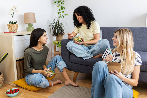 Three women are enjoying a healthy meal together, sharing salads and conversation in a comfortable living room setting. Female friendship and youth concept