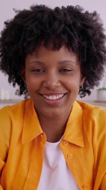 Portrait of cheerful young African American woman dressed in casual clothes, sitting at a table and smiling at the camera. Relaxed lifestyle, positive emotions, leisure and home atmosphere concept.