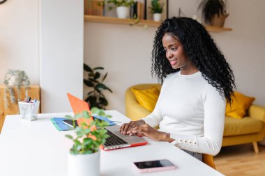 Genç Latin Amerikalı siyahi kız öğrenci evde bilgisayar üzerinde çalışıyor. Modern apartmandaki masada dizüstü bilgisayar kullanan neşeli etnik kadın portresi. Eğitim ve iş konsepti.
