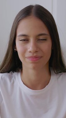 Evde neşeli bir şekilde oturan gülümseyen genç kızın dikey HD videosu. Kameraya gülümseyen mutlu gençlik portresi. 