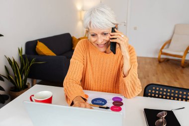 Evde dizüstü bilgisayarla çalışan, telefonda konuşan ve iş planı hakkında not alan kıdemli iş kadını. Girişimciler ve iş konsepti