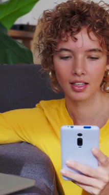 Genç beyaz kadının dizüstü bilgisayar ve akıllı telefon kullanarak evde birden fazla iş yaptığı dikey HD videolar. Teknoloji ve iletişim konsepti. 