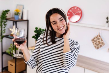 Mutlu güzel bir kadın internet müziğinin keyfini çıkarırken kablosuz kulaklıkla kırmızı şarap içiyor ve evde mutfakta şarkı söylüyor.