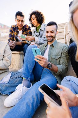 Üniversite basamaklarında birlikte oturan genç öğrenciler cep telefonlarını kullanırken ve iletişim anlarını paylaşırken. Arkadaşlık ve eğitim yaşam tarzı.