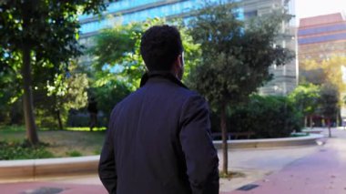 Güneşli bir günde modern cam binalarla çevrili yeşil bir şehir parkında yürüyen zarif iş adamının arka görüntüsü. Çağdaş finans bölgesinde kendine güvenen bir profesyonel gezinti.