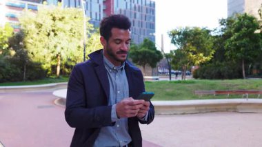 Akıllı telefonunu kontrol ederken modern bir şehir parkında yürüyen kendinden emin genç ve başarılı bir işadamı arka planda çağdaş bir binayla işe giderken birden fazla iş yapıyor.