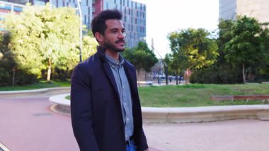 Sakallı ve kıvırcık saçlı yakışıklı Afro-Amerikan bir adam ağaçlar ve binalarla dolu modern bir şehir parkında güvenle yürüyor, güneşli bir günün tadını çıkarıyor.