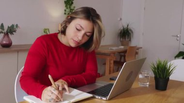 Masasında oturan, not defterine not alan ve uzaktan kumandalı işi için dizüstü bilgisayar kullanan genç bir kadın rahat ve modern bir ofis ortamında görevlerine odaklandı. İş veya eğitim kavramı