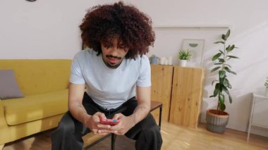 Genç yetişkin Latin Amerikalı adam, modern oturma odasında otururken cep telefonu uygulamasında video oyunu oynamaktan zevk alıyor, boş zamanlarında odak ve mutluluk gösteriyor.