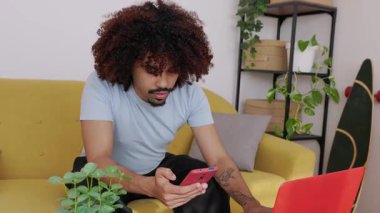 Bilgisayarında çalışan kıvırcık saçlı, odaklanmış genç Afrikalı Amerikalı adam aynı anda akıllı telefonunu kontrol ediyor, modern oturma odasındaki sarı bir kanepede oturuyor.
