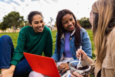 Çeşitli gülümseyen kız öğrenciler yeşil çimlerde oturuyor, dizüstü bilgisayar kullanıyor, gülüyor, dışarıda atıştırıyor, arkadaşlığımızı kutluyor ve birlikte öğreniyoruz.