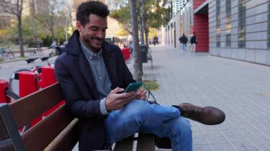 Genç, yetişkin, Latin Amerikalı bir adam bankta akıllı telefon kullanarak gülümsüyor. Sakin profesyonel modern kentsel alanda dijital iletişimin keyfini çıkarıyor. Teknoloji ve yaşam tarzı kavramı.