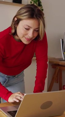 Genç bayan girişimcinin dikey HD videosu ahşap bir masada durup daktilo yazarken modern, rahat bir ev ofisinden çevrimiçi işlerini yönetirken aynı zamanda gülümsüyor ve verimli hissediyordu.