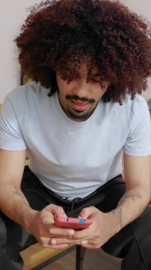 Modern oturma odasında oturup boş zamanlarında odak ve mutluluk gösteren, cep telefonu uygulamasında video oyunu oynayan genç Latin Amerikalı adamın dikey HD videosu.