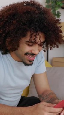 Oturma odasındaki sarı koltukta oturan genç bir adamın akıllı telefonunu kullanarak sosyal medyaya mesajlaşırken ve gezinirken çekilmiş dikey HD videosu.