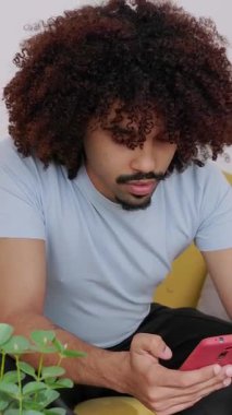 Bilgisayarında çalışan kıvırcık saçlı, modern oturma odasındaki sarı koltukta oturan ve aynı zamanda akıllı telefonunu kontrol eden Afro-Amerikan bir adamın dikey HD videosu.