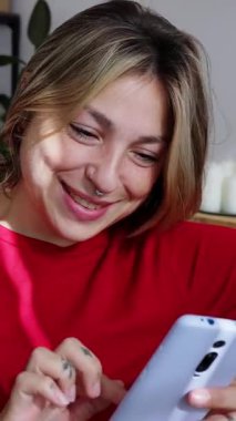 Samimi bir oturma odasında sarı bir koltukta oturan, gülümseyen ve gülen genç bir kadının cep telefonuyla mesajlaşırken ve kaydırırken evinde rahatlatıcı bir anın keyfini çıkardığı dikey videosu.