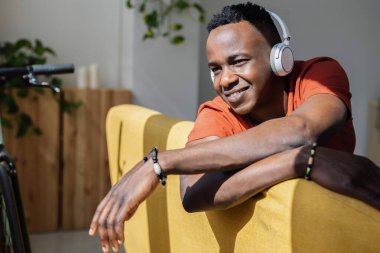 Rahat Afrikalı adam kanepede müzik dinliyor pencereden gün ışığının tadını çıkarıyor. Ilık güneş ışığı yüzünü aydınlatıyor. Huzurlu ve rahat bir atmosfer yaratıyor. Rahatlık ve farkındalık..