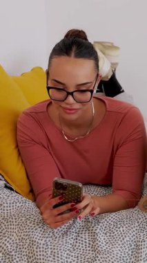 Yatakta rahatlarken cep telefonuna göz atan genç bir kadının dikey HD videosu. Genç kız birlikte rahat bir dijital anın tadını çıkarıyor. Teknoloji ve sosyal medya kavramı