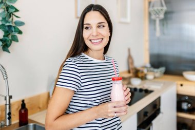 Genç, mutlu bir kadın, elinde pipetle taze meyve kokteyli tutarken gülümsüyor. Çağdaş bir ev mutfağında duruyor ve sağlıklı bir yaşam tarzı ve refahı üzerine odaklanıyor.