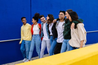 Bir grup neşeli genç öğrenci mavi bir duvarın önünde birlikte yürüyüp birlikte vakit geçiriyorlar.