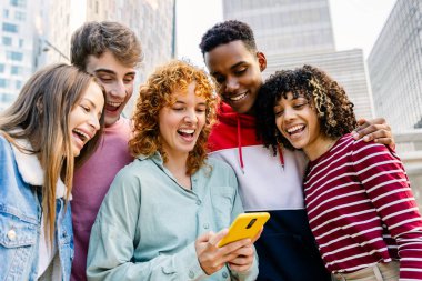 Bir şehir caddesinde gülen ve bir akıllı telefonu paylaşan bir grup genç, spontane bağlantı, arkadaşlık ve modern dijital birlikteliğin tadını çıkarıyorlar.