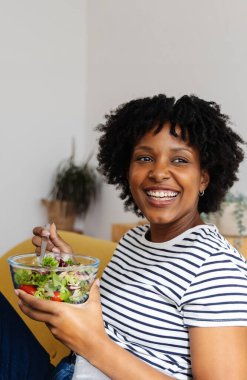 Mutlu genç Afrikalı Amerikalı kadın evdeki kanepede oturmuş sağlıklı bir salatanın tadını çıkarıyor.