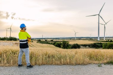 Rüzgar tarlasında çalışan mühendis, rüzgar türbinleriyle dolu bir alanda duruyor ve temiz yenilenebilir enerji üretiyor. Yenilenebilir enerji ve endüstriyel işgücü kavramı